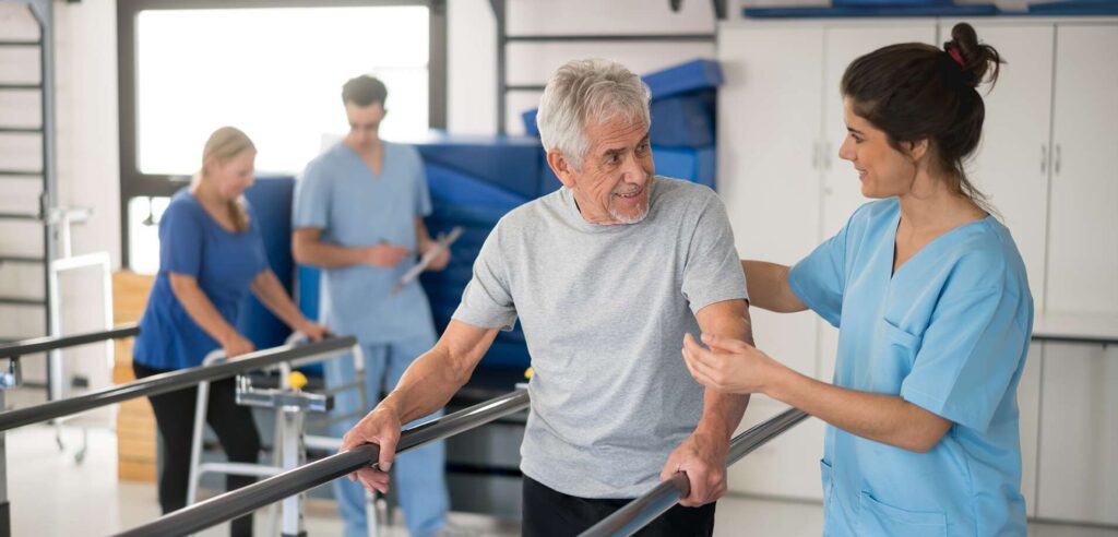 Cheerful senior man using parallel bars to walk and physiotherapist smiling at him happy - Skilled Nursing